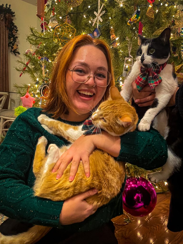 Kennedy, an orange/yellow-haired woman with square glasses, cradles an orange cat like a baby.