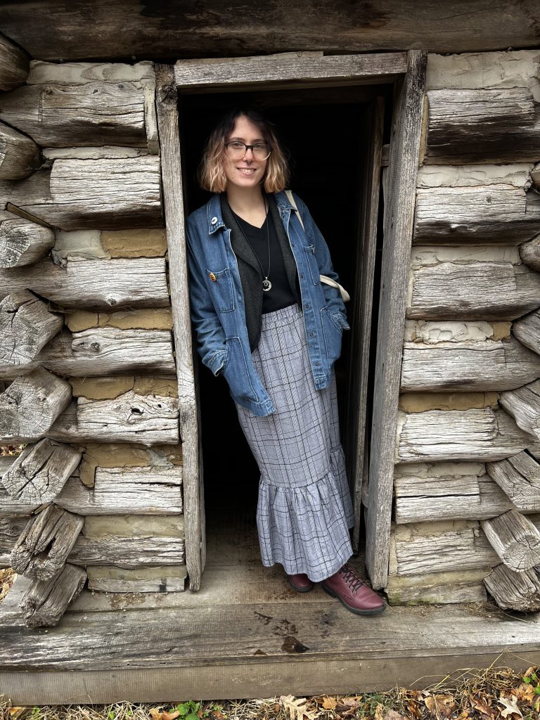 Spring, a person with black and blonde hair and glasses, leans against the frame of a log cabin door.