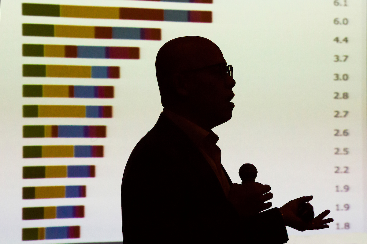 A speaker is silhouetted against a bar graph showing several declining values.