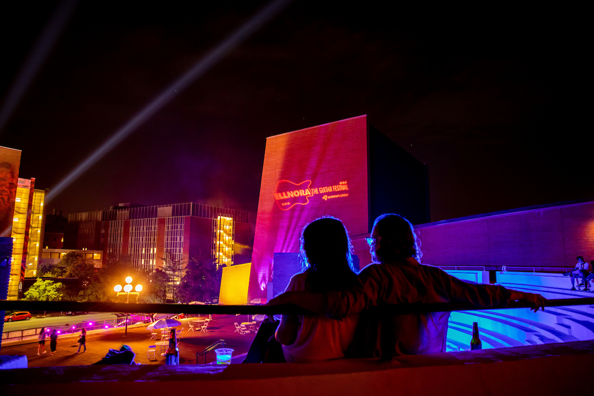 Two blue-silhouetted figures sit at the edge of the outdoor stage in front of an orange Ellnora Festival light projected on the wall of Krannert Center.