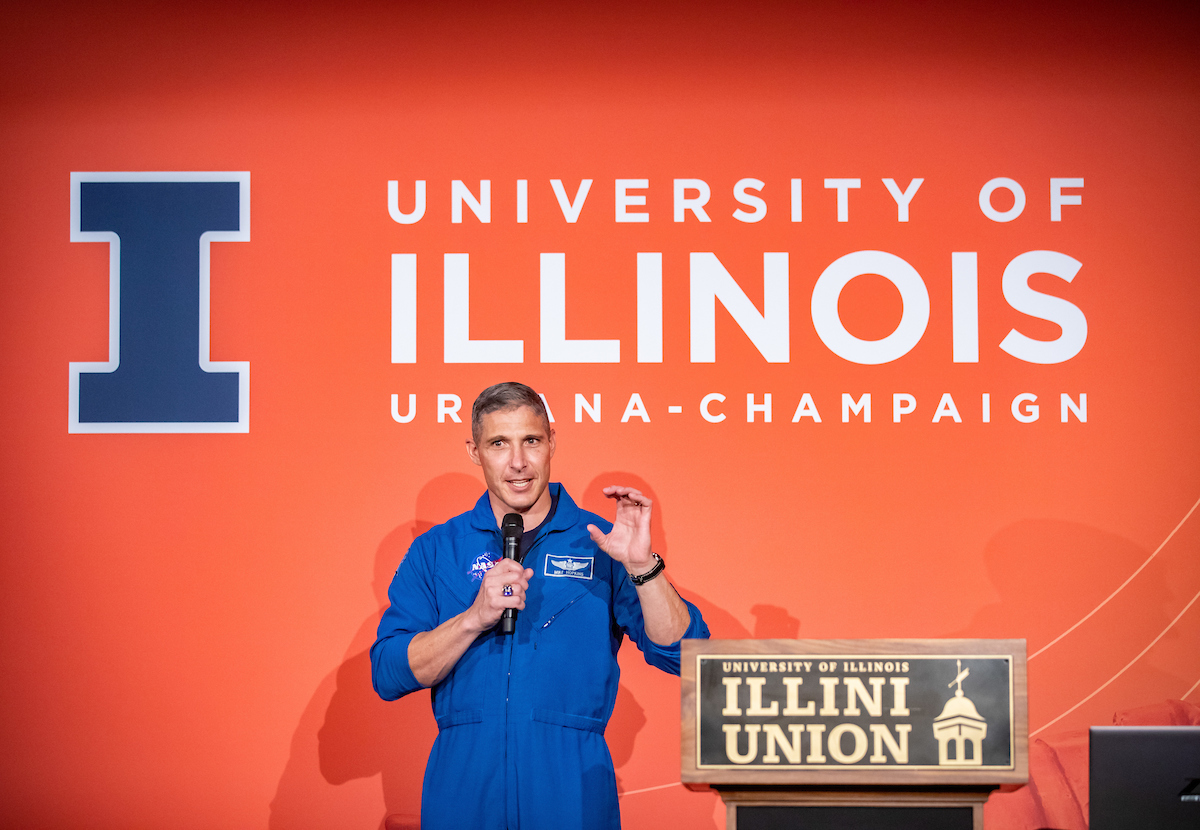 Col. Mike Hopkins stands in front of an orange screen and beside an Illini Union podium